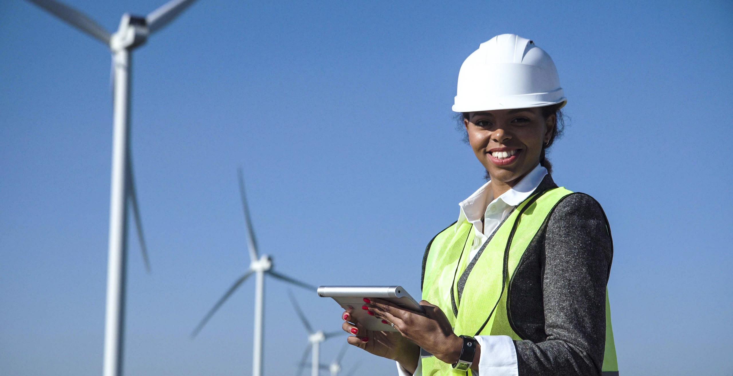 Female engineer wearing hard hat standing with digital tablet against wind turbine on sunny day and looking at camera smilingly.