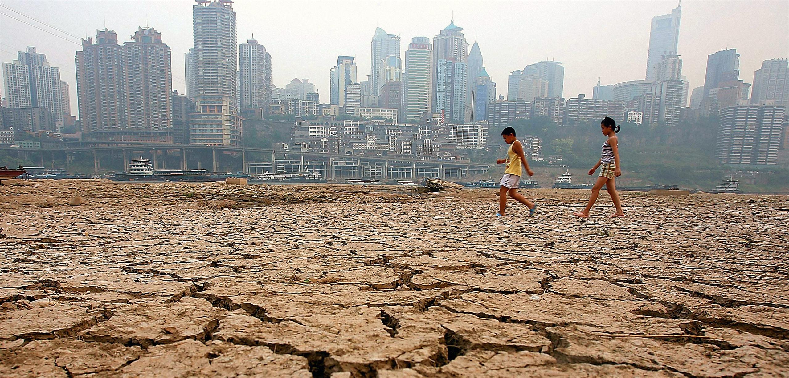 Two young children walking across cracked, dry land with a city skyline behind.