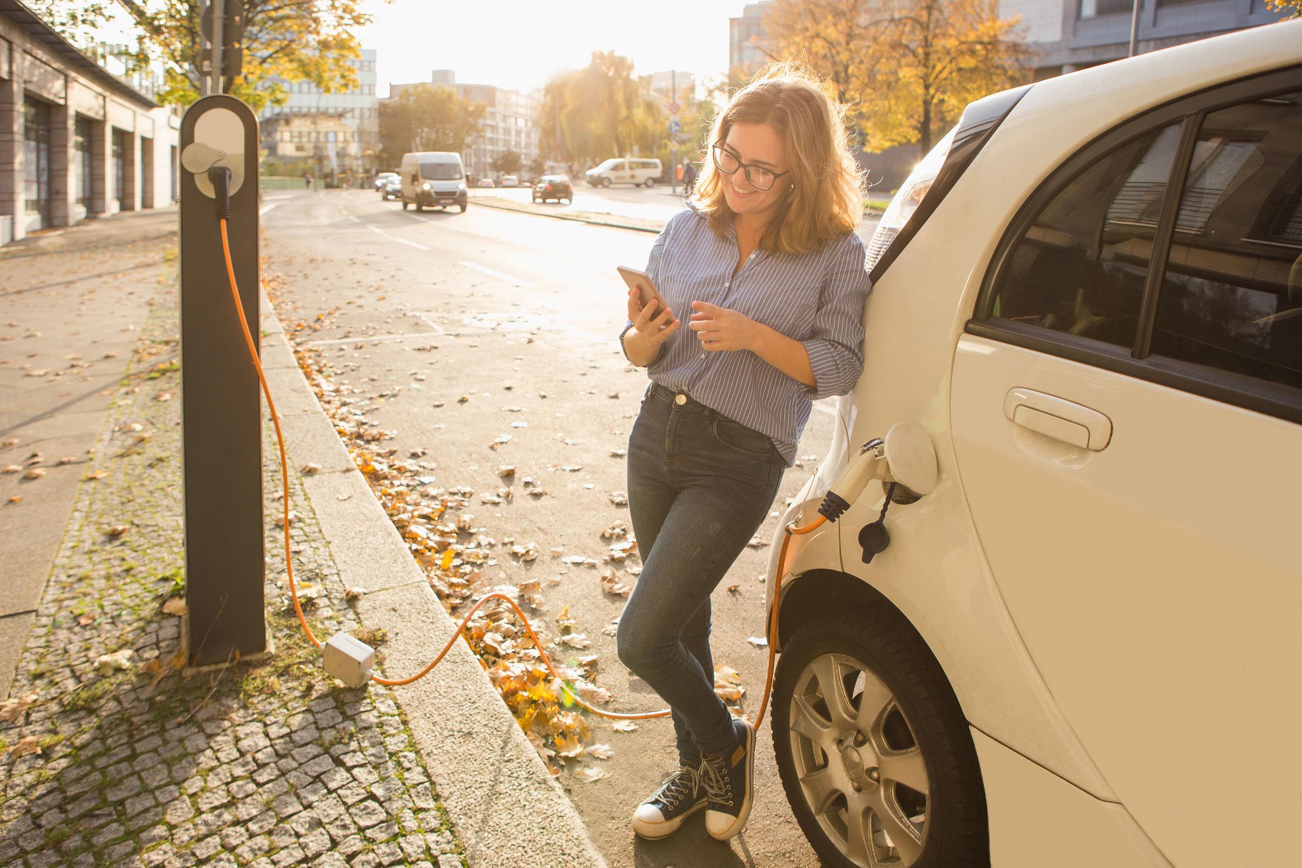 A young woman using her mobile phone whilst waiting for her electric car to charge.