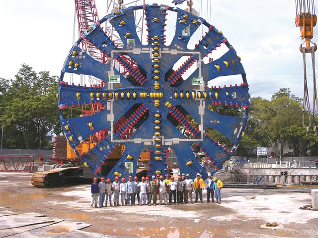 Shot of giant tunnel boring machine dwarfing workers standing in front of it.